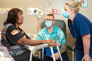 An Aboriginal woman sits on the side of a bed holding the top of a walking frame as she talks with a male and female health professional wearing surgical masks.