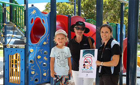 A young boy stands with two woman in front of colourful playground equipment. One woman holds a certificate that reads Please do not smoke near our playgrounds.