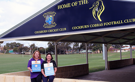 Two women holding signs that read 'Proud to be Smoke Free'. The women are seated underneath a sign that reads 'Home of the Cockburn Cricket Club and Cockburn Cobras Football Club'